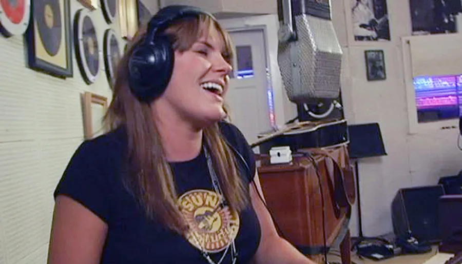A person is smiling and laughing while wearing headphones in a room with musical equipment and gold record plaques on the wall.