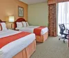 The image shows a tidily kept hotel room with a large bed a desk with a chair a television and colorful curtains