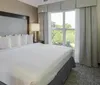 A neatly made king-sized bed is the centerpiece of a modern hotel room with large windows offering a view of greenery outside