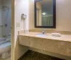 The image shows a neutrally decorated hotel room with a large bed an attached bathroom wall-mounted lights and a small desk area