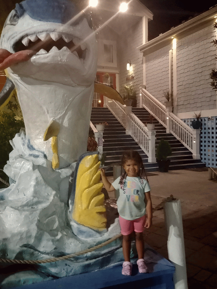 A child stands smiling next to a large shark statue outside a building at night.