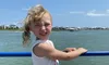 A child squints in the bright sunlight while smiling and holding onto a blue railing on a boat with a backdrop of water and houses.