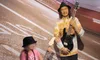A child wearing a pink shirt and a hat plays a guitar next to a wax figure of a male guitarist, in an exhibit with a road-themed backdrop.