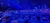 A crowd of people is seated in a theater, some of them talking, surrounded by blue lighting.