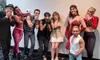 A diverse group of people in costumes and stage makeup, ranging from gothic to sporty styles, are posing together with some covering their mouths with their hands.
