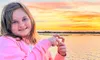 A girl in a pink sweatshirt poses happily near a body of water with a vibrant sunset in the background.