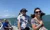A group of people are enjoying a sunny day on a boat, with a smiling woman in sunglasses standing at the railing as the wind blows her hair, and a man in a patterned shirt and hat beside her.