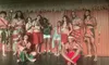 A group of people dressed in traditional Polynesian attire are posing on a stage, smiling and making hand gestures.