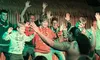 A group of people is participating in a lively dance performance led by a person with tattoos and traditional attire, raising their hands enthusiastically.