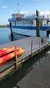 A large blue and white boat is docked near a pier with stacked kayaks and a coiled hose in the foreground.