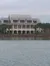 A large multi-story house with a metal roof is situated along a calm waterfront, surrounded by palm trees.