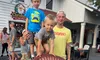 A man stands behind two children climbing on a colorful dinosaur statue outside a 
