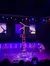 A performer balances on one hand atop a stack of chairs during a stage show, with musicians playing on either side and an audience member capturing the moment on their smartphone.