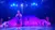 A performer dressed in a colorful outfit stands on a brightly lit stage holding a hat, with several other performers visible in the background.