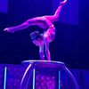 A performer exhibits remarkable flexibility and balance in a contortion act on a circular platform under blue stage lighting.