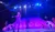 A performer in a purple outfit is dancing or skating on a brightly lit stage with vibrant pink and blue lighting, surrounded by geometric structures and other performers in the background.