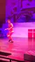 A performer in a red outfit is energetically playing a diabolo on stage with a colorful backdrop depicting a snowy village scene.