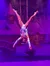 A performer in colorful attire is suspended upside down on ropes against a snowy village backdrop.