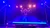 A performer in vibrant lighting is balancing on a slackline suspended above a stage.