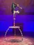 A performer is executing an acrobatic handstand on a curved candy-cane-striped pole attached to a table, against a backdrop suggesting a snowy village at night.