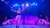 A performer is posing with arms outstretched on a stage under blue and purple lighting, with several other individuals in the background.