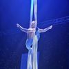 A performer is suspended mid-air, striking a dramatic pose with outstretched arms, wrapped in long fabric from an aerial silk performance, against a starry backdrop.