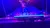 A performer is suspended upside down on a brightly lit aerial wire in a vibrant, colorful stage setting.