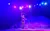 A performer on a brightly lit stage is skillfully twirling a lighted hoop while standing in a confident pose under vibrant pink and purple lighting.