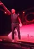 A performer on stage dramatically points into the audience under red lighting, with another person in the background.