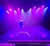A performer stands on a circular apparatus under vibrant purple lighting on a stage.