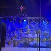 A performer walks blindfolded on a high circular apparatus with a snowy village backdrop.