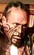 A performer wearing traditional Maori attire, including a feathered cape and facial paint, is energetically holding a spear above his head.