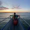 A person in a tie-dye shirt sits at the bow of a boat, gazing towards a serene sunset over the water.