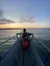 A person in a tie-dye shirt sits at the bow of a boat, gazing towards a serene sunset over the water.