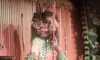 A person in cultural attire holds a carved staff during a performance on a stage with thatched decor elements in the background.