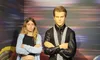 A person is posing with arms crossed next to a life-like wax figure dressed in a formal black suit, set against a background with a colorful motion blur effect.