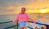 A person stands smiling at the camera on a boat with a beautiful sunset in the background.