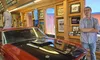 A person stands with arms crossed next to a classic car in a room filled with automotive memorabilia.