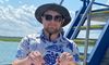 A person wearing sunglasses and a hat is holding a crab on a boat with a maritime-themed shirt, under clear blue skies.
