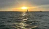 A sailboat glides on the ocean as the sun sets on the horizon, casting golden reflections on the water's surface.