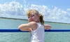 A smiling child leans on a blue railing aboard a boat, with a scenic view of water and clouds in the background.