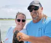 A smiling man and woman are posing on a boat with a freshly caught fish, with fishing rods and the water visible in the background.