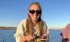 A smiling person in sunglasses is holding a crab on a boat with water and shoreline in the background.