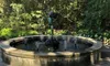 A stone fountain with a statue of a winged figure holding a bow is situated among lush greenery and trees.
