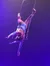 A trapeze artist performs an acrobatic pose mid-air under purple stage lighting.