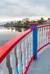 A vibrant red, white, and blue railing curves around the deck of a boat with a scenic view of a river and tree-lined shore.