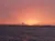 A vibrant sunset casts warm orange and pink hues over the ocean, with a silhouette of distant buildings on the horizon.