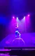 An acrobat performs a balancing act on a high platform, illuminated by purple and blue stage lights, holding a bow.