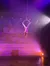 An aerial performer in a white costume executes a move on a hoop suspended from the ceiling, with a wintry village backdrop and colorful stage lighting.