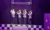 Four performers in silver suits sing on stage beneath a sign reading 